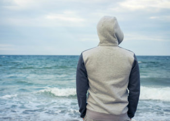 A man stands on the beach in a tracksuit with a hood and looking at the sea. Cloudy weather, rear view.